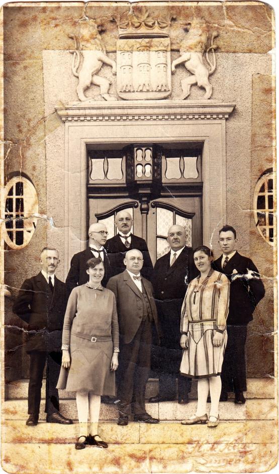 Die schwarz-weiß Fotografie zeigt eine Gruppe von Menschen, darunter sechs Männer und zwei Frauen, die auf einer Treppe vor dem Eingang des Bürgermeisterhauses stehen. 